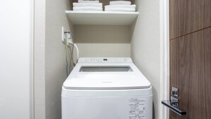 A compact, white front-loading washing machine in a small laundry room with a wooden door and a shelf above holding folded towels. - 1 min walk from sta./Excellent access to central (Osaka)