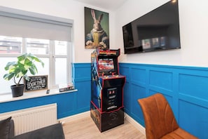 Ein Zimmer mit Spielkonsole, Blumentopf, Wandfernseher und einem Fenster mit Jalousien.