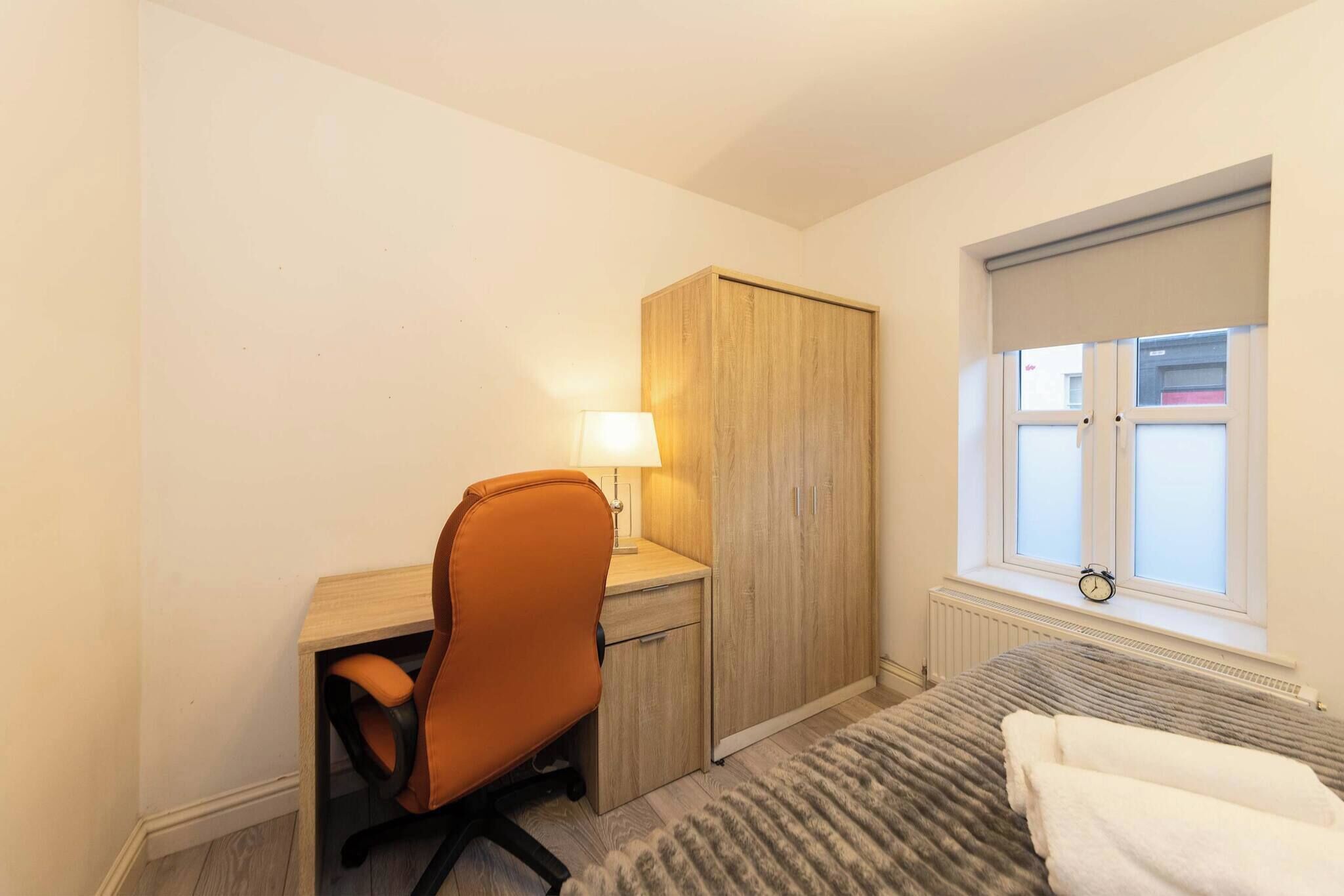Ein kleiner, ordentlicher Raum mit Schreibtisch aus Holz, einem orangefarbenen Sessel, einem Holzschrank, einem Fenster mit Aussicht und einer Heizung.