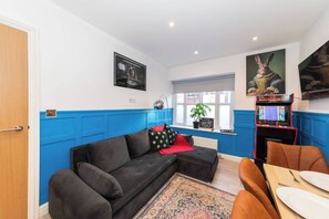 A room with a dark grey sofa, a red throw pillow, a blue wall, a poster, and a gaming console. - Ibex House Cozy Modern 2-bed Flat Free Parking (Reading)