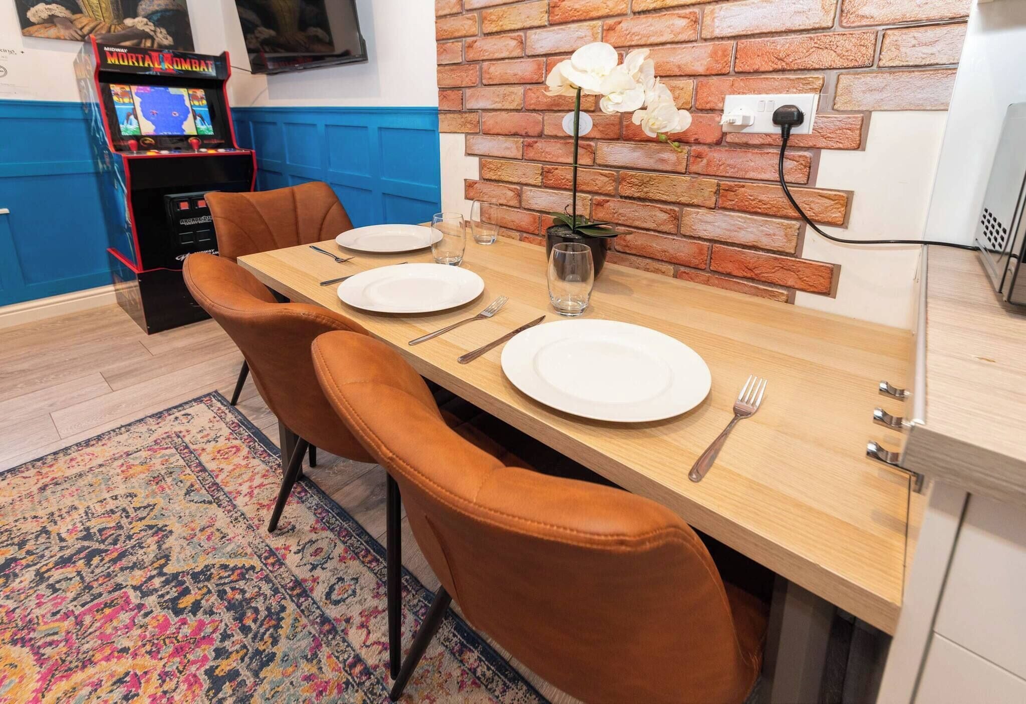 Un coin repas avec un mur en briques, une table en bois dressée pour deux personnes et, au fond, une machine de jeux.