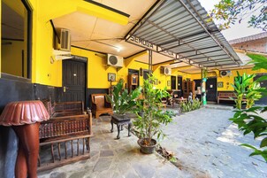 Un patio con zona de estar techada, plantas en macetas y un edificio con fachada amarilla.