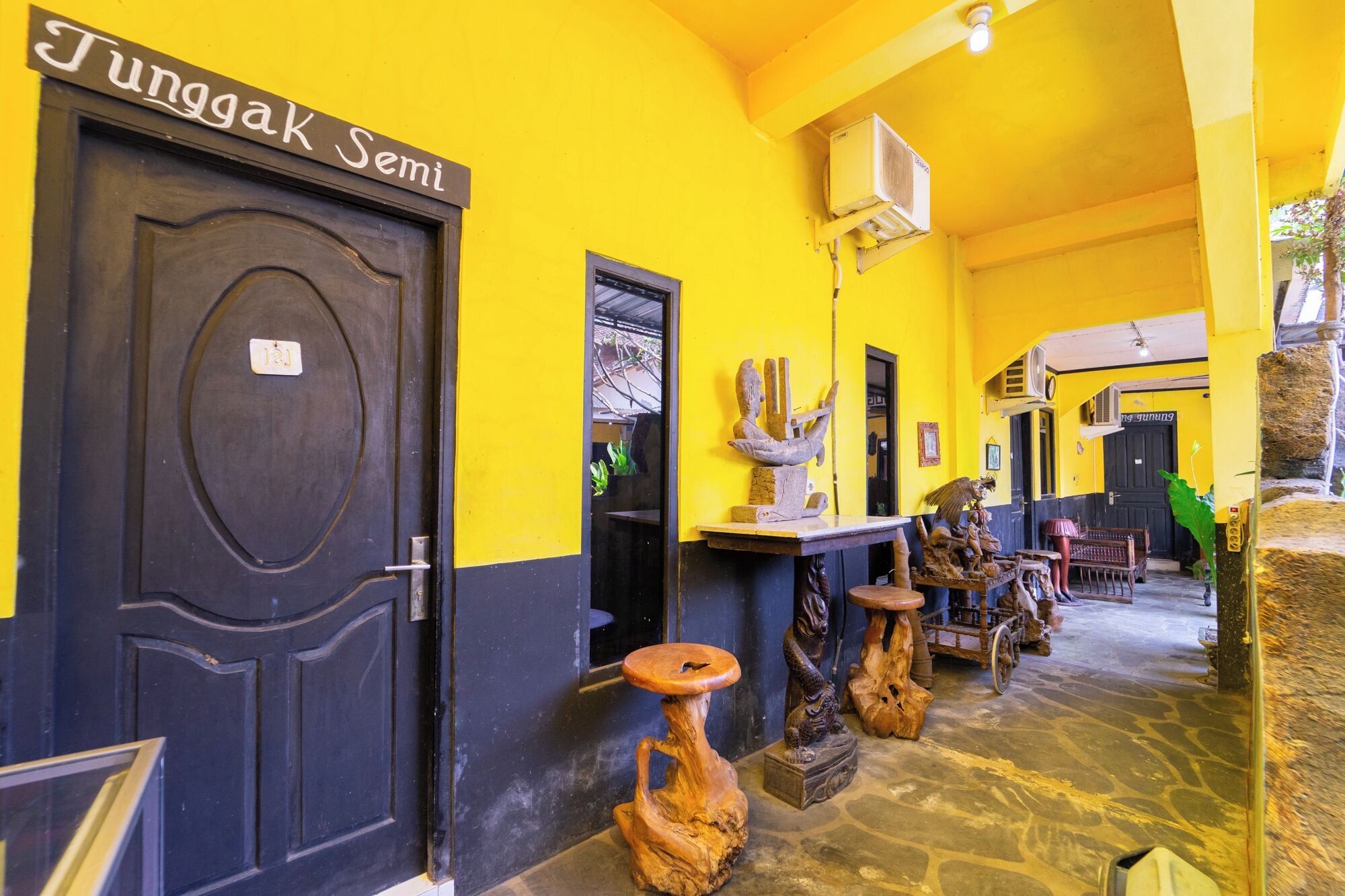 A hallway with a dark door labeled 'Jungak Semi,' a yellow wall, and various wooden sculptures.