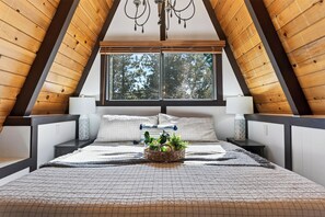 Ein Schlafzimmer mit schrägem Dach, einem großen Fenster, einem Bett mit gemustertem Überwurf, zwei Nachttischen mit Lampen und einer Pendelleuchte.