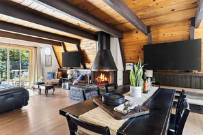 A spacious living area with a fireplace, wooden ceiling beams, a large flat-screen TV, and a dining table with chairs. - A-Frame | Hot Tub Deck | Fireplace | Trails, Golf & Marina | A Big Bear View (Big Bear Lake)