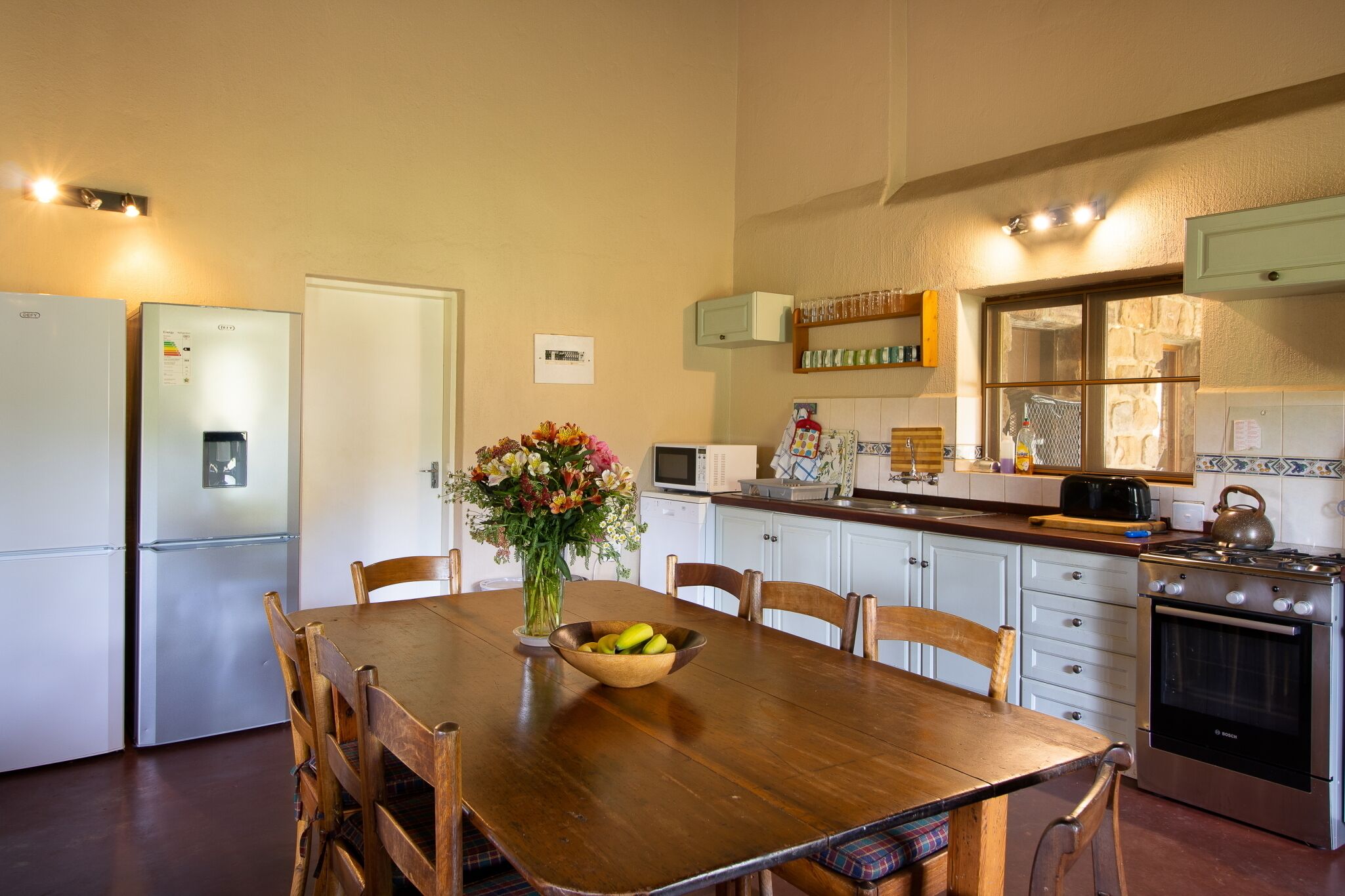 A kitchen with a wooden dining table, chairs, a refrigerator, a microwave, a stove, and a flower vase.
