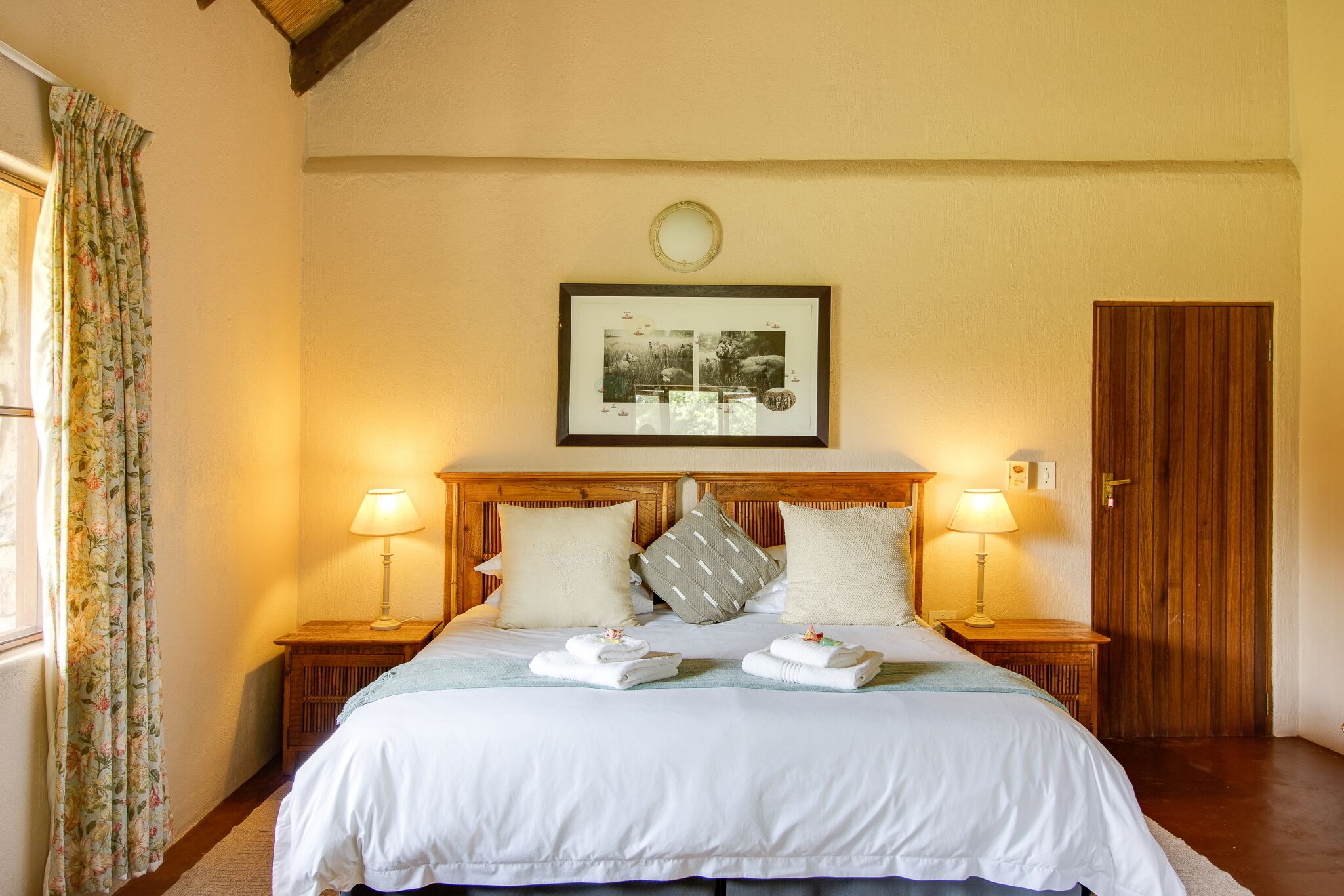 Een hotelkamer met een groot bed, twee bedlampen, een lijst met een afbeelding boven het bed en een houten deur.