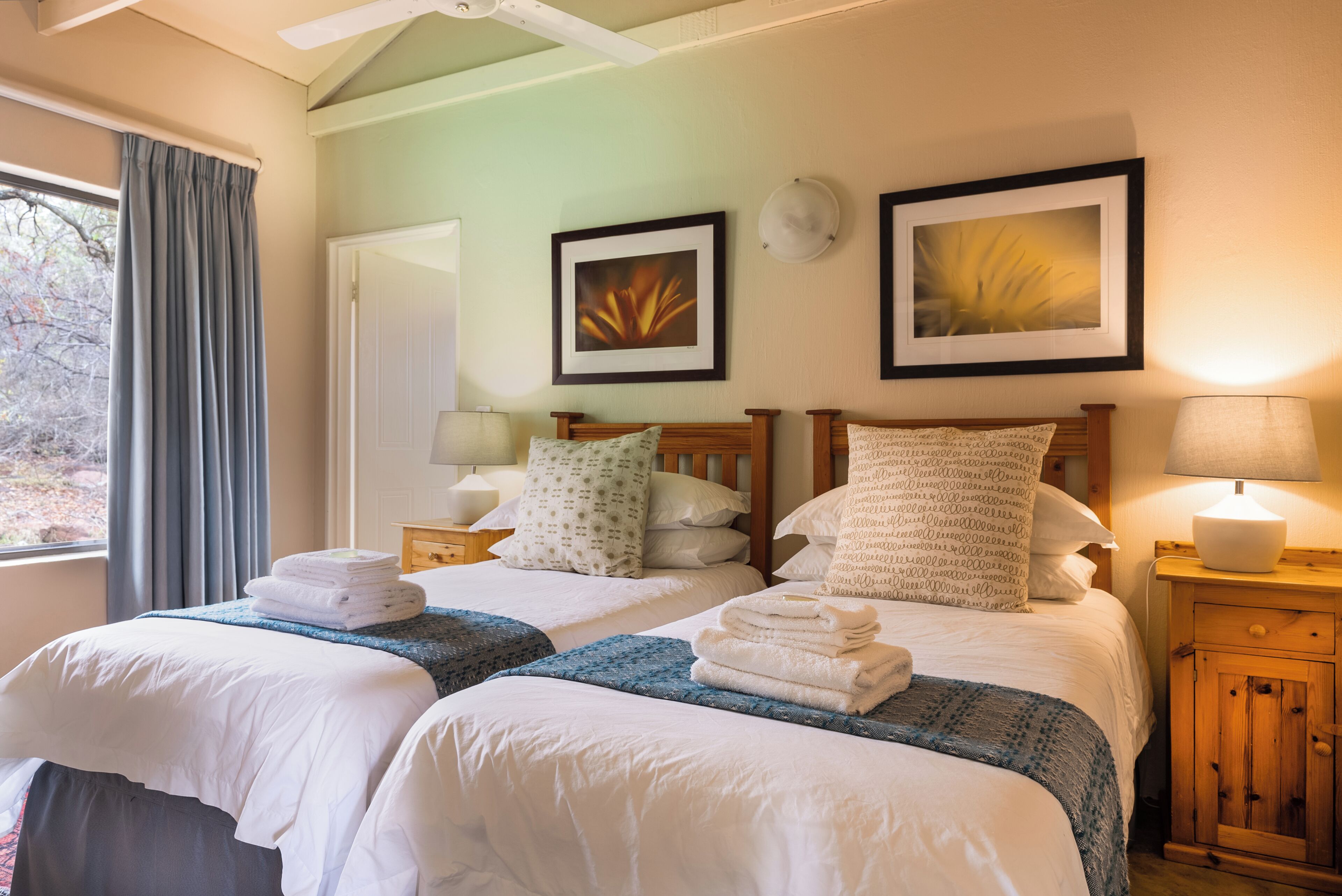 Quarto de hotel com duas camas, janela com cortinas, ventilador de teto e duas imagens emolduradas na parede.