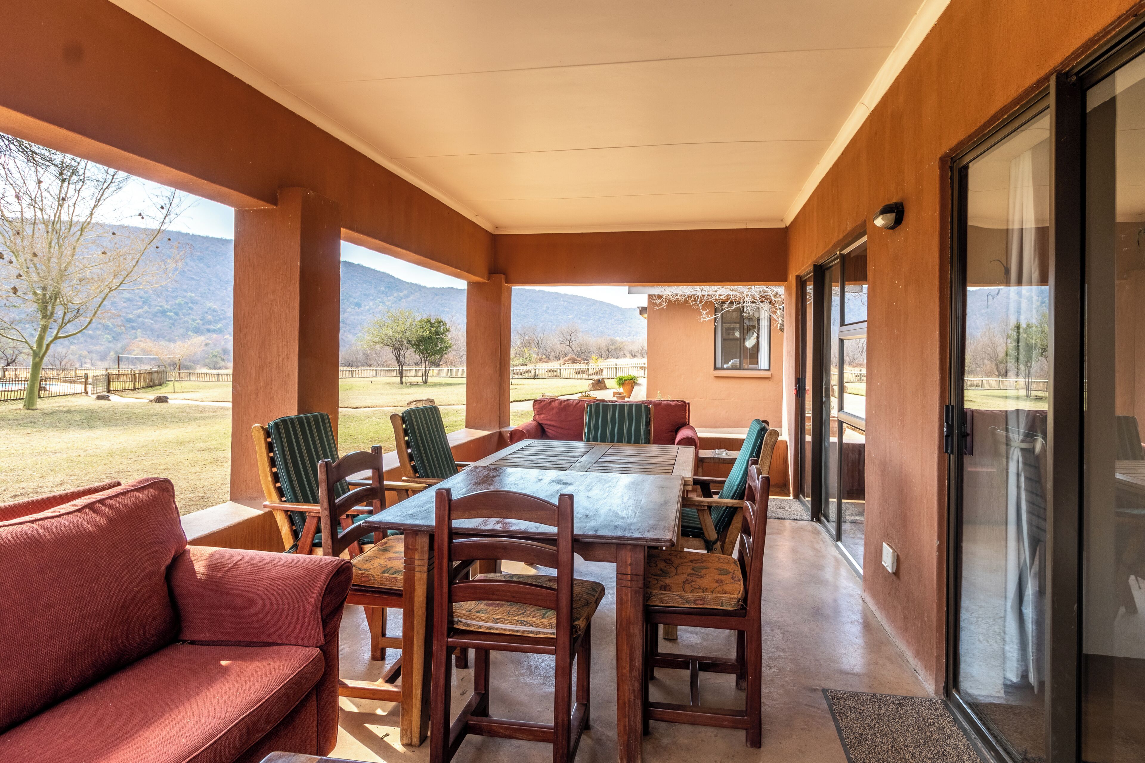 A covered patio area with a wooden table and chairs, a red sofa, and a view of a mountainous landscape.