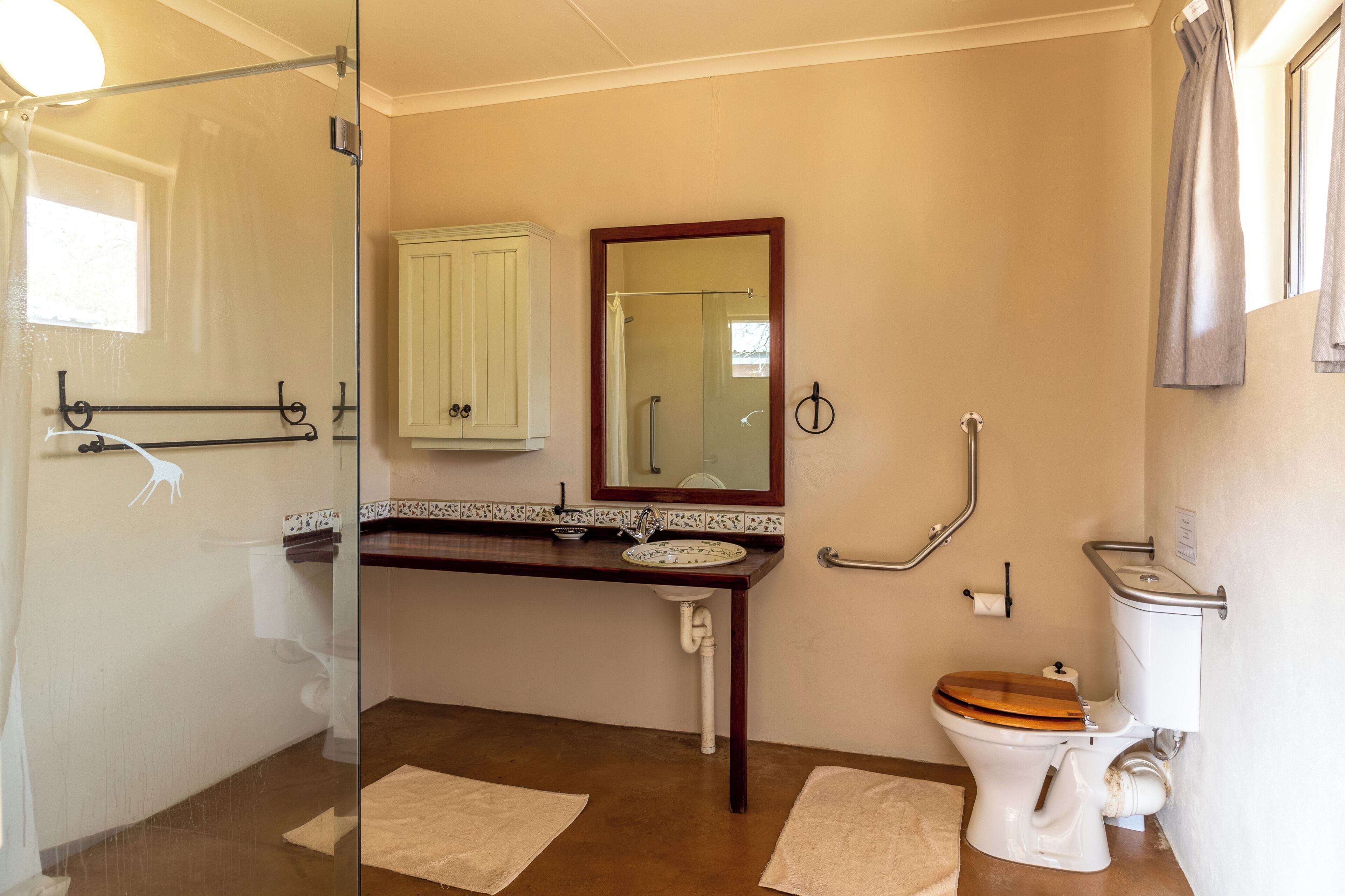 A bathroom with a glass shower enclosure, a wooden toilet, a sink with a mirror above it, and two towels on the floor.