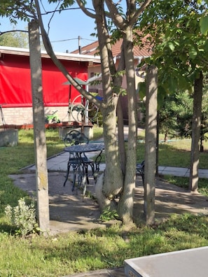A patio area with a table and chairs, surrounded by trees and a red building in the background.