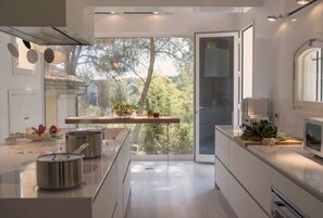 A modern kitchen with a central island, stainless steel pots, a glass door leading to a balcony, and a view of trees. - Elegant 7 Bedroom Villa, Pool and Panoramic Sea Views (Costa Saracena-Castelluccio)