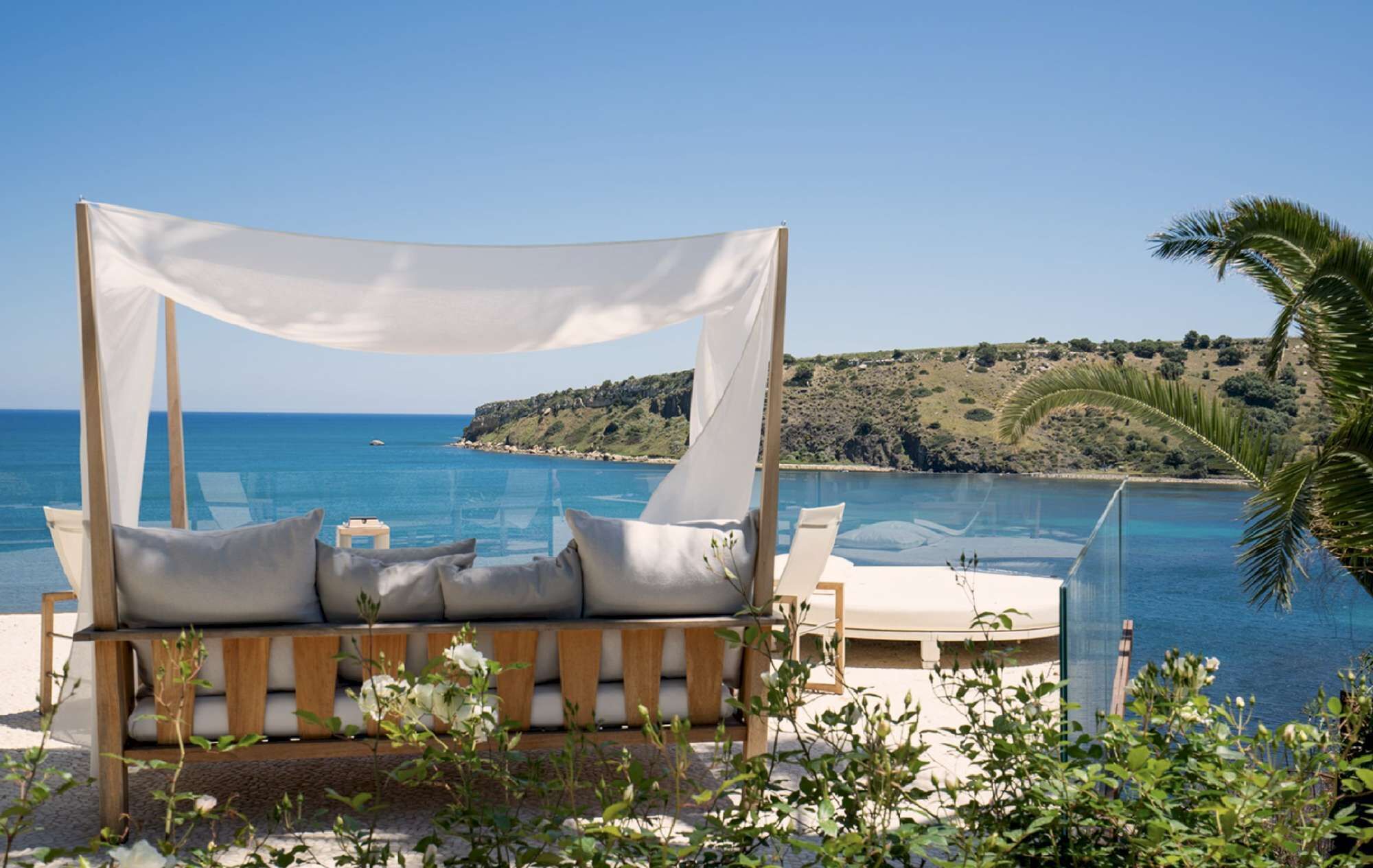 Eine Strandlounge mit Baldachin, einem Pool und Blick auf das Meer und eine hügelige Landschaft.