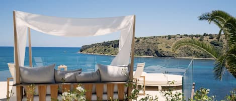 Eine Strandlounge mit Baldachin, einem Pool und Blick auf das Meer und eine hügelige Landschaft.