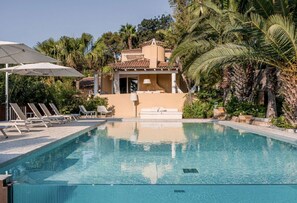 A clear swimming pool with lounge chairs and umbrellas, surrounded by palm trees and a building with a tiled roof. - Elegant 7 Bedroom Villa, Pool and Panoramic Sea Views (Costa Saracena-Castelluccio)