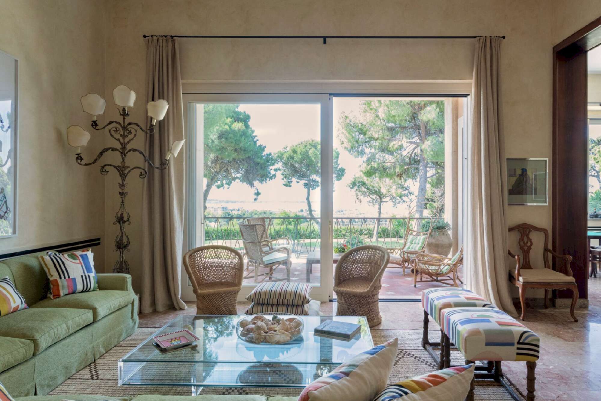 A spacious living room with a glass coffee table, wicker chairs, a sofa, and a view of a garden through large sliding doors.