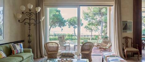 A spacious living room with a glass coffee table, wicker chairs, a sofa, and a view of a garden through large sliding doors.