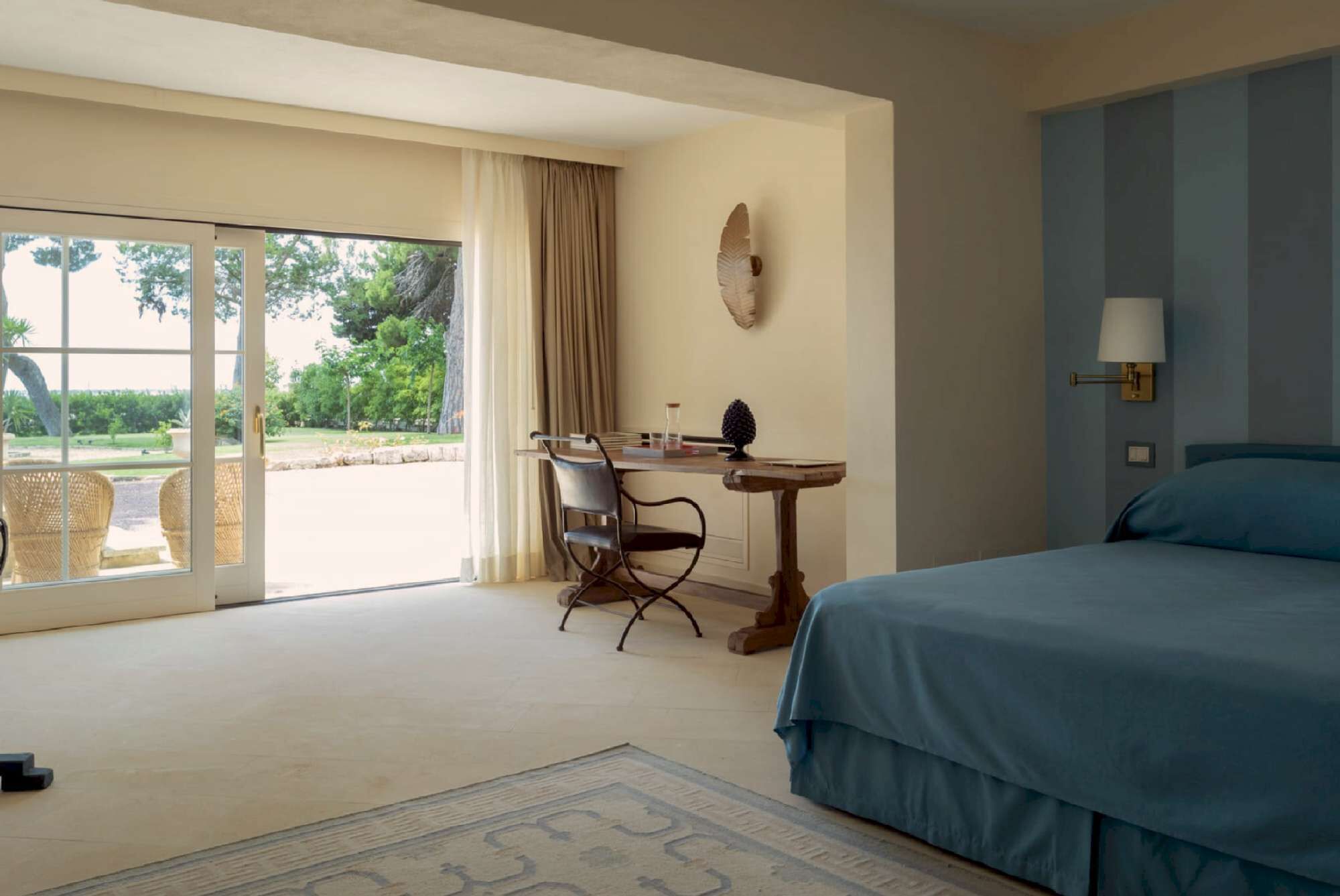 A hotel room with a bed, a desk, a chair, a lamp, and a view of the outdoors.
