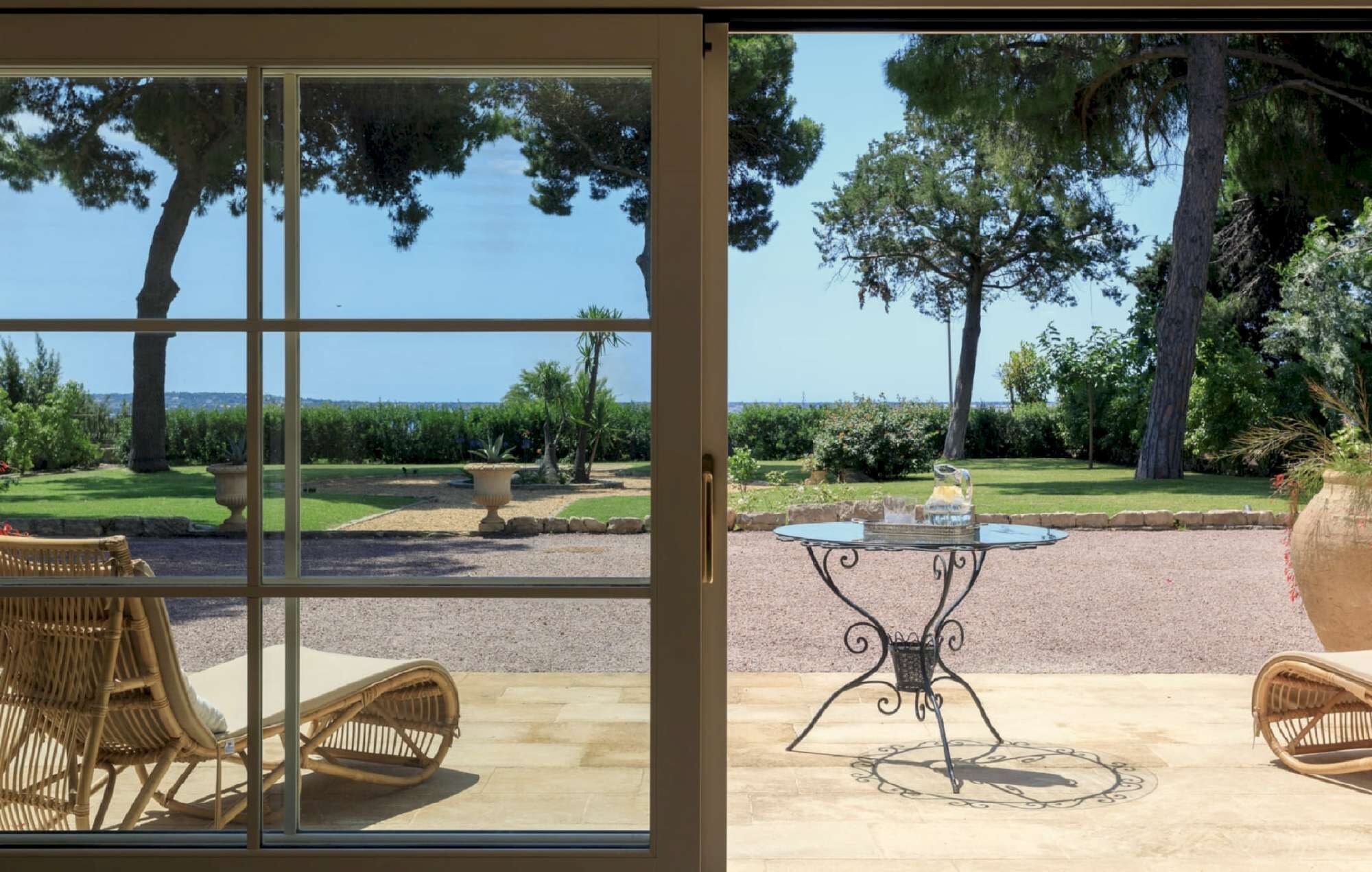 A patio with wicker furniture, a metal table, and a view of a landscaped garden with trees and a clear sky.