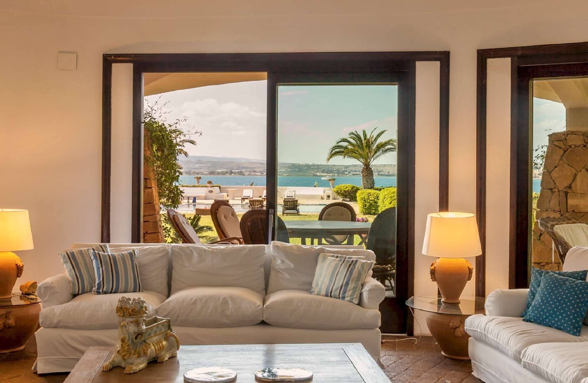 A living room with a sofa, a coffee table, and two lamps. There is a view of a pool and palm trees through a glass door.
