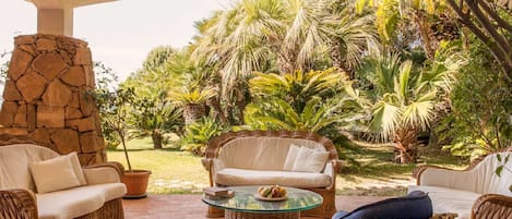 Un patio al aire libre con mobiliario de mimbre, un pilar de piedra y exuberante vegetación tropical.