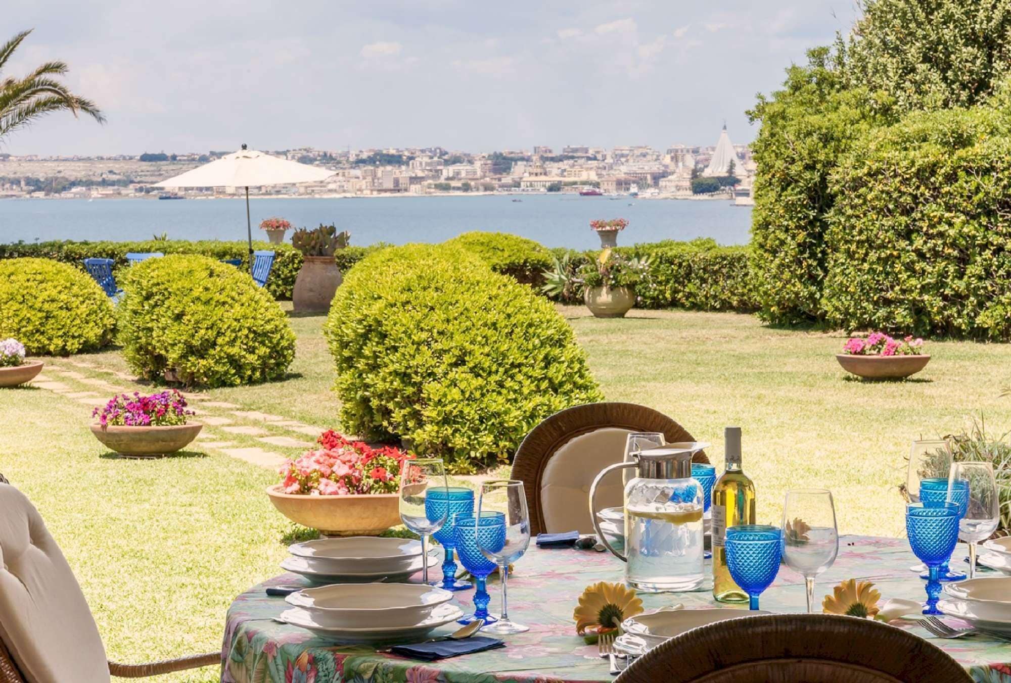 Un espace de restauration en plein air offrant une vue sur la mer et le paysage urbain, avec des tables disposées avec soin, agrémentées de verres bleus et de compositions florales.