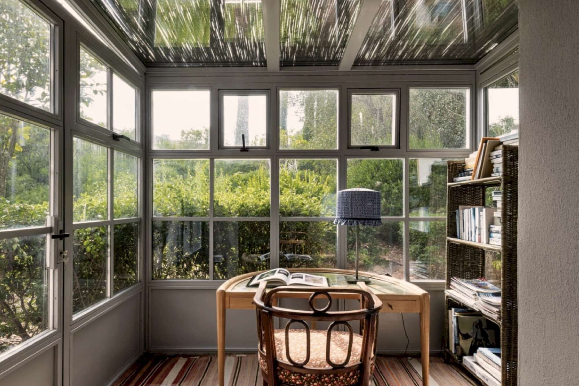 A room with a large windowed bay window, a wooden chair, a table with a book, and a bookshelf.