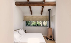 Ein Schlafzimmer mit Bett, einem Fenster mit Vorhang, Holzbalkendecke und Kaminofen.