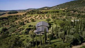 A house with a sloped roof surrounded by a well-maintained garden with tall trees and a hilly landscape in the background. - Large Orbetello Villa with Private Pool (Orbetello)