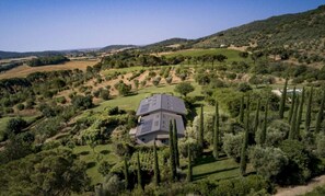 A house with a sloped roof surrounded by a well-maintained garden with tall trees and a hilly landscape in the background. - Large Orbetello Villa with Private Pool (Orbetello)