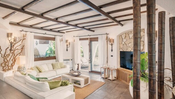 Ein geräumiges Wohnzimmer mit hoher Decke, einer weißen Sofagarnitur, einem Couchtisch und einem Fernseher.