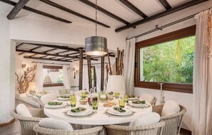 A dining area with a round table set for four, surrounded by wicker chairs, a hanging light fixture, and a view of greenery through a window. - Sardinia Dream Getaway: 4BR Villa with Pool & Garden (Santa Margherita di Pula)