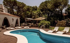 Une piscine dotée de chaises longues et d’un espace salon couvert, le tout entouré d’arbres et d’un mur en pierre.