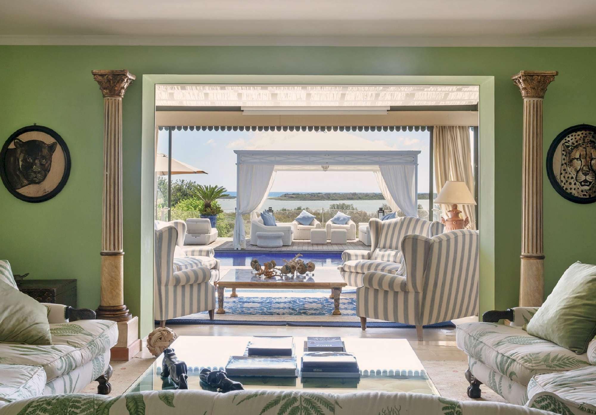 A spacious living room with a view of a pool and the sea, furnished with striped armchairs and a coffee table.