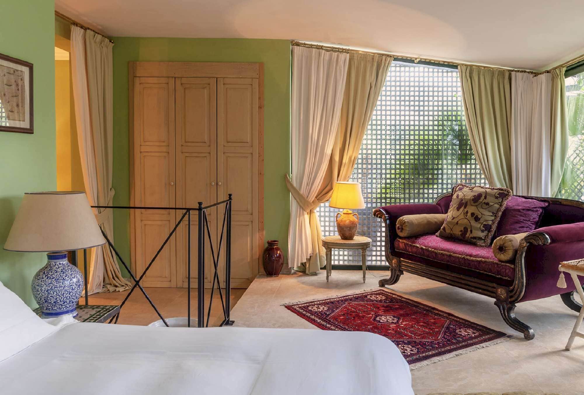 Une pièce comprenant un lit, un canapé violet, une table avec une lampe, une fenêtre avec des rideaux et une porte.