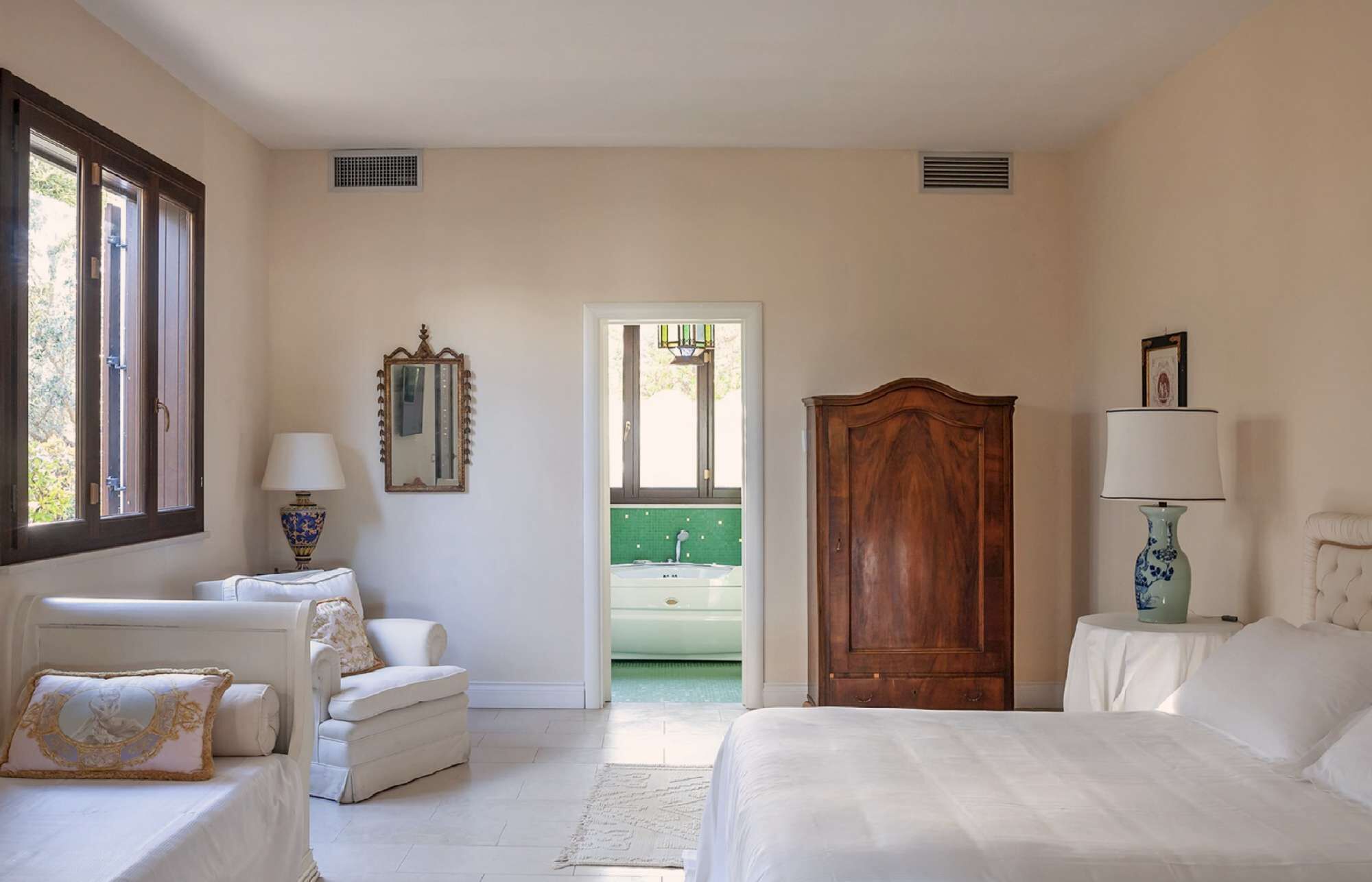 A bedroom with a bed, two chairs, a wooden wardrobe, a mirror, and a window.