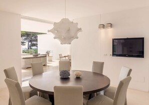 Un espace repas moderne avec une table ronde, six chaises, un luminaire suspendu original et un téléviseur à écran plat fixé au mur.