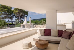 Ein moderner Außenbereich zum Sitzen mit einer Couch und einem runden Hocker, mit Blick auf eine malerische Landschaft mit Bäumen und einer entfernten Hügelkette.