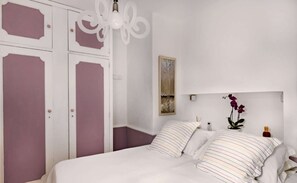 Ein Schlafzimmer mit einem Bett, zwei Kissen, einem Nachttisch, einem kleinen Tisch, einer Wandlampe und einer Tür mit dekorativer Verkleidung.
