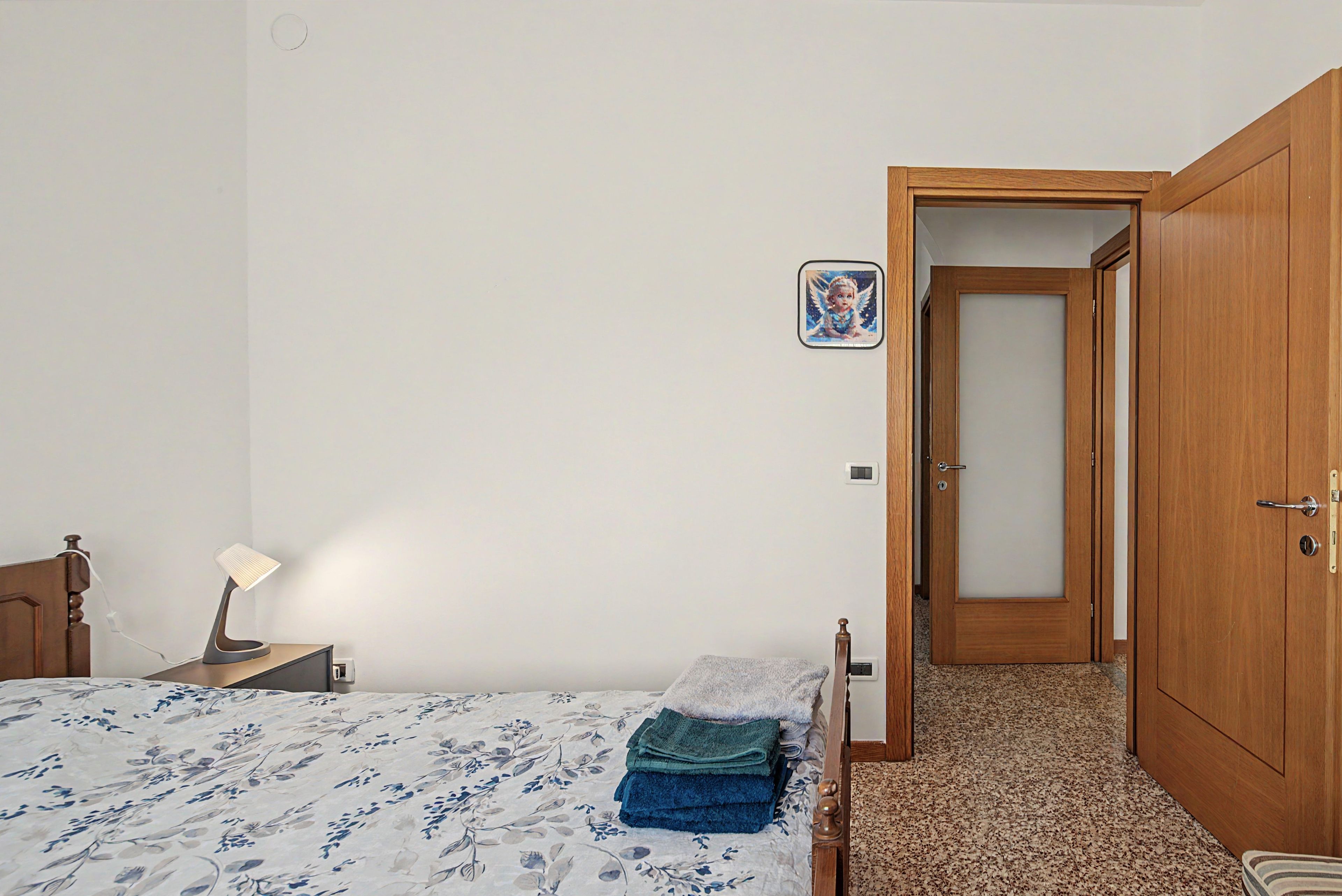 A bedroom with a bed, a bedside table with a lamp, a door, and a wall-mounted picture.