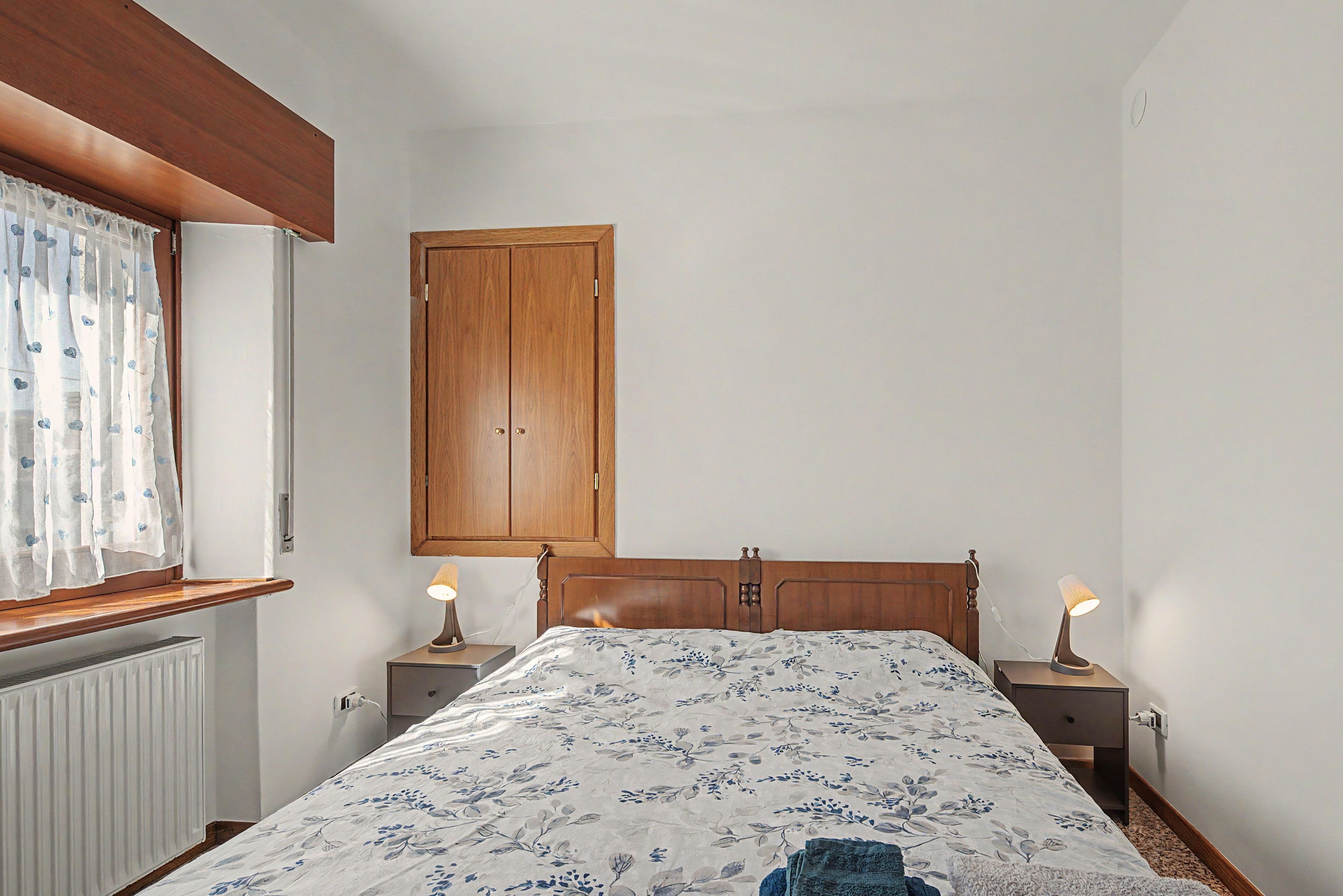 Un dormitorio con cama, dos mesitas de noche con lámparas, un armario de madera y una ventana con cortina.