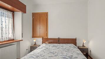 Un dormitorio con cama, dos mesitas de noche con lámparas, un armario de madera y una ventana con cortina.