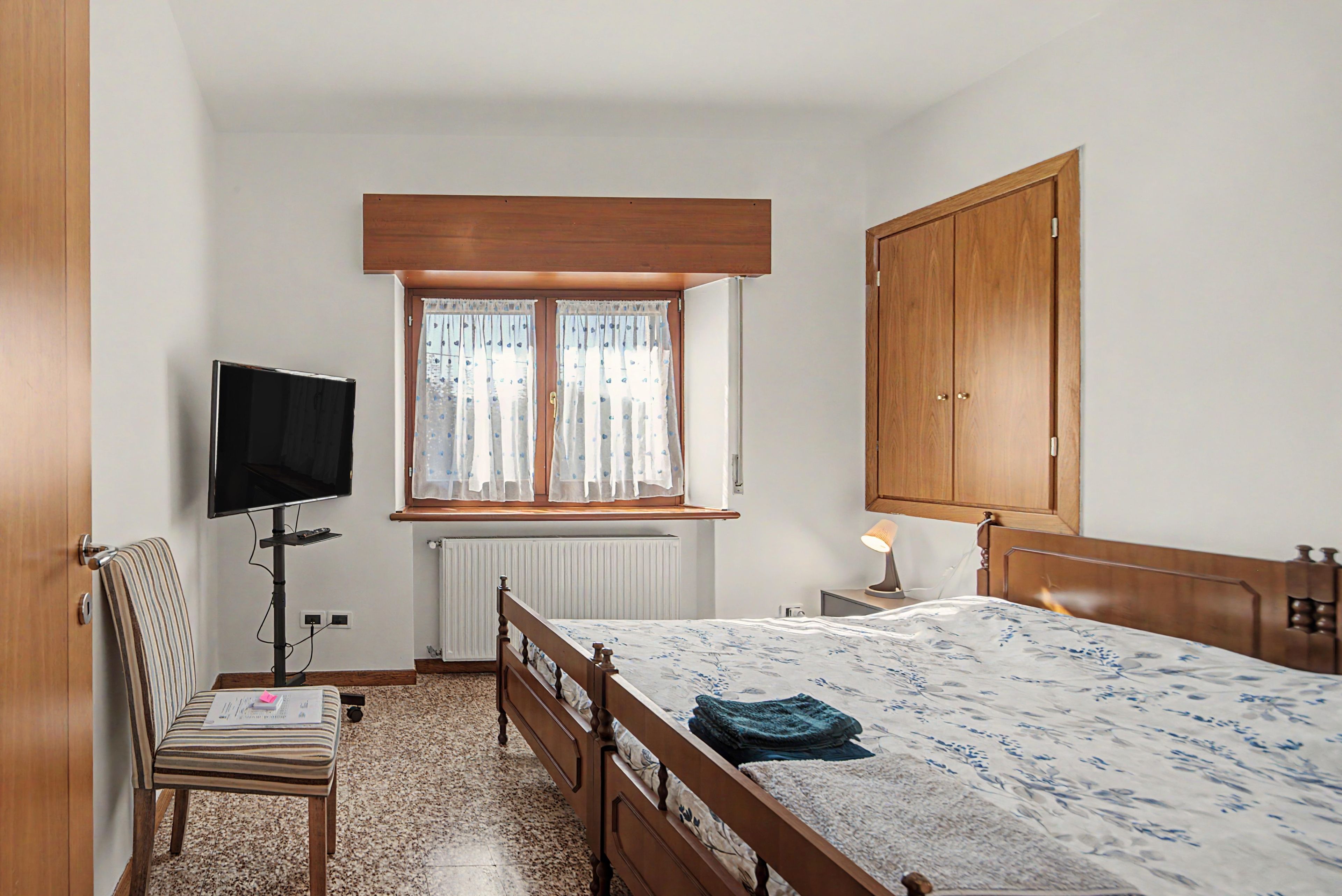 Un dormitorio con cama, una silla, televisión, ventana con cortinas, radiador y armario.