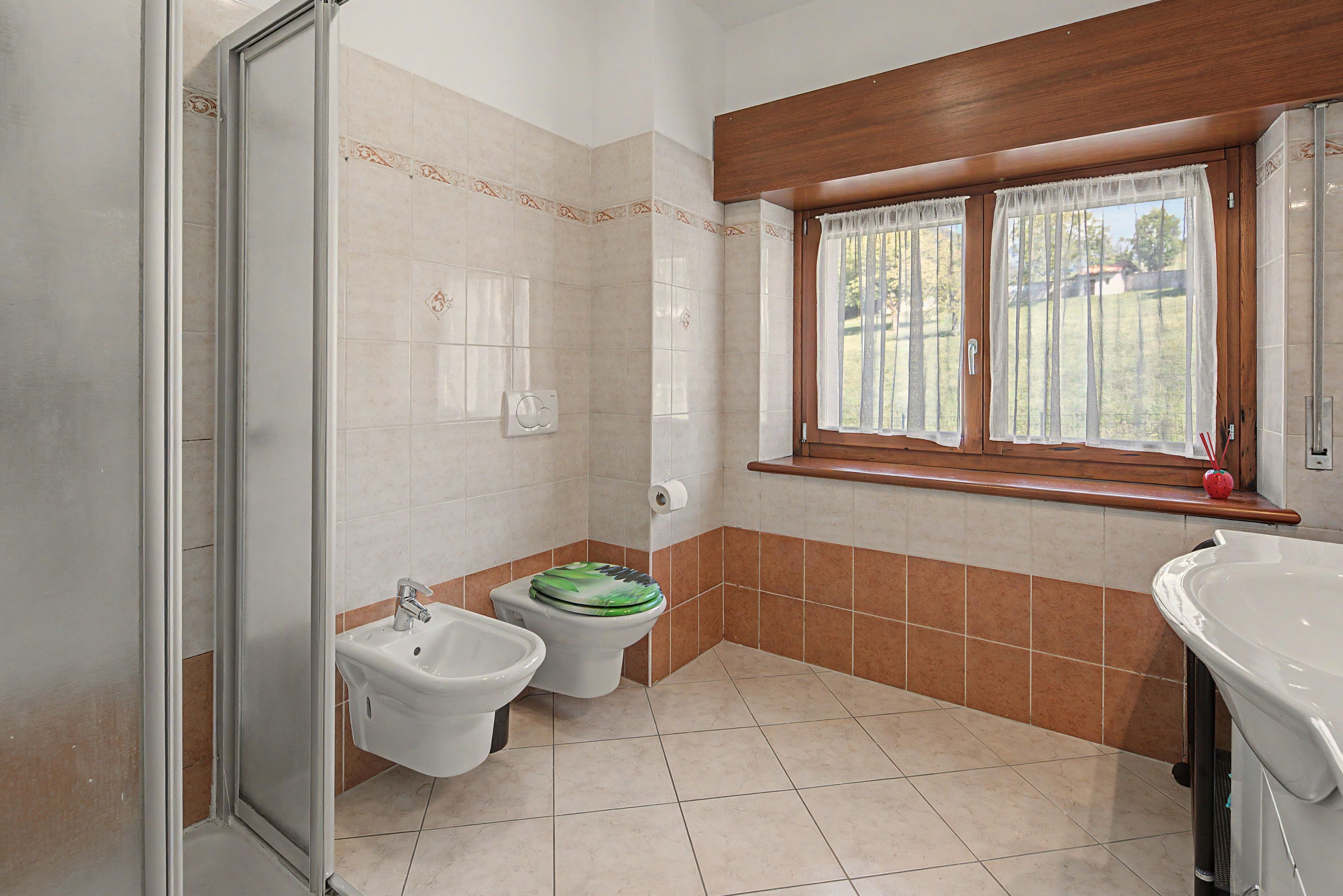 A bathroom with a shower, two bidets, a toilet, a window with curtains, and a sink.