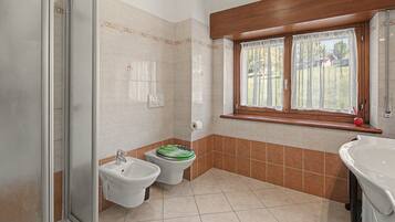 A bathroom with a shower, two bidets, a toilet, a window with curtains, and a sink.