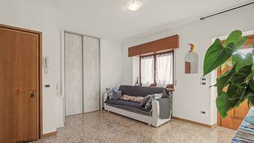 Una habitación con sofá, puerta de madera, ventana con cortinas y una planta grande.