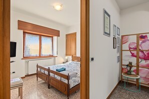 Un dormitorio con una cama de madera, una ventana con cortinas, un radiador, una mesita de noche y una pared decorada con diplomas enmarcados.
