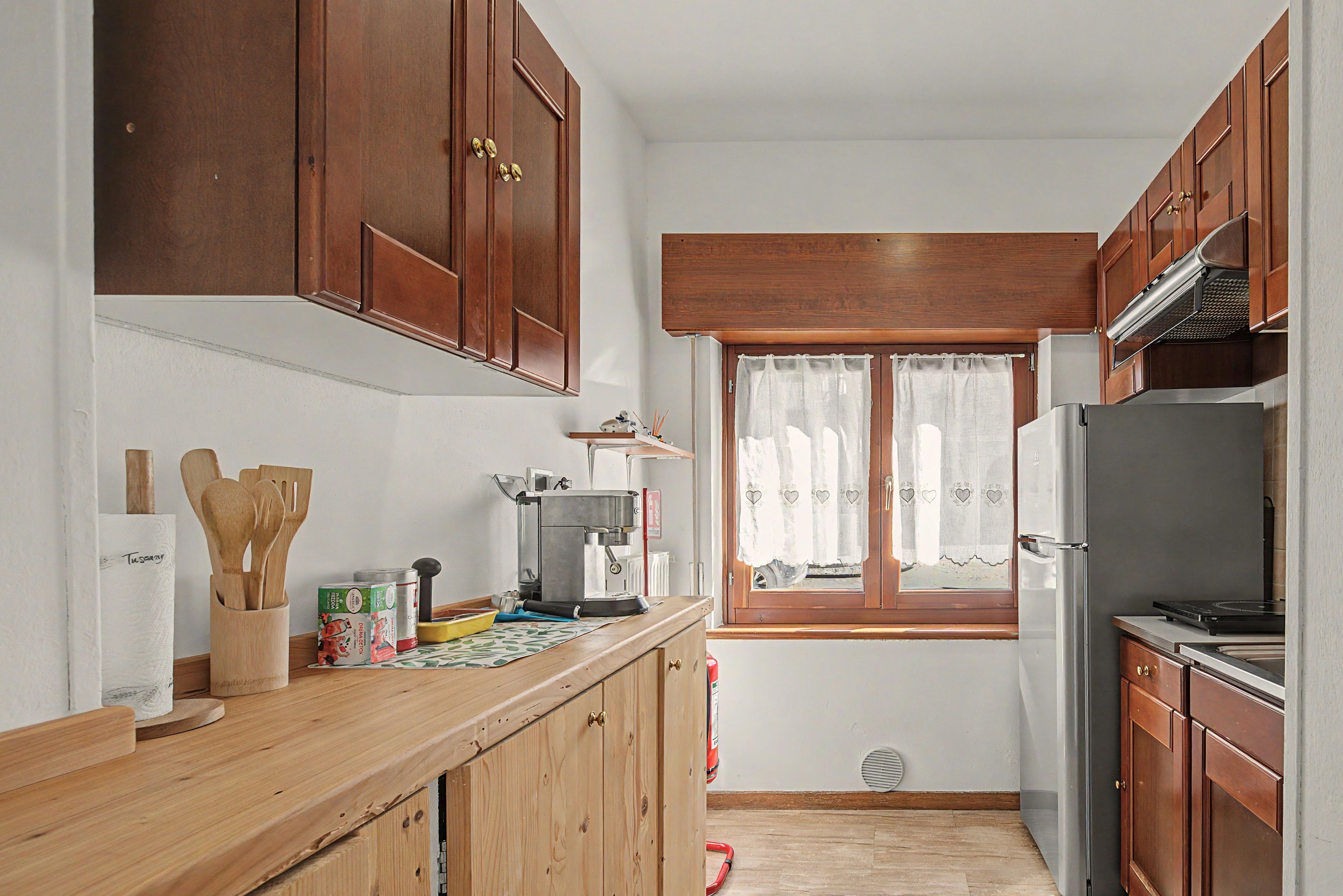 A kitchen with wooden cabinets, a coffee machine, a refrigerator, and a window with curtains.