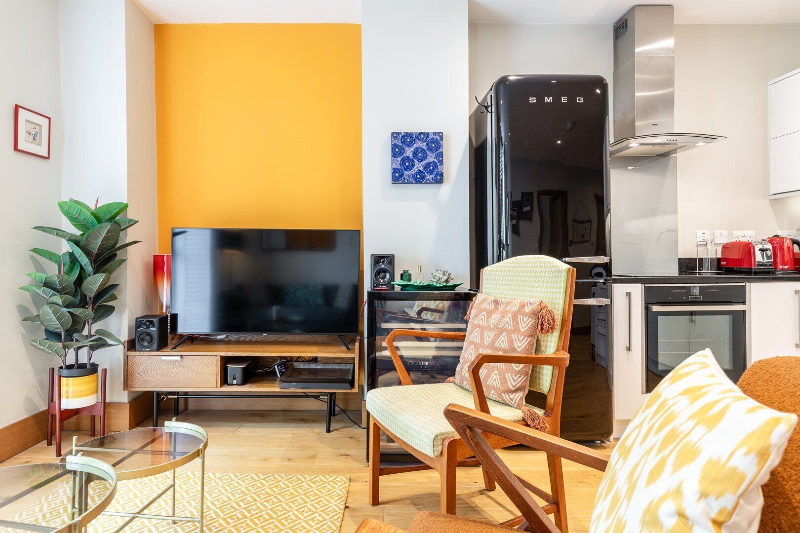 A modern living room with a flat-screen TV, a wooden chair, a glass coffee table, and a kitchen area with a refrigerator and oven.
