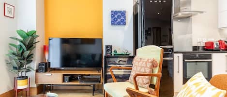 A modern living room with a flat-screen TV, a wooden chair, a glass coffee table, and a kitchen area with a refrigerator and oven.